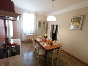 Un coin repas est aménagé avec une table en bois pour quatre personnes, entourée de chaises blanches. On y trouve également un coin cuisine avec une cuisinière et une bouilloire posées sur le plan de travail. Une fenêtre ornée de rideaux blancs laisse entrer la lumière naturelle dans la pièce.