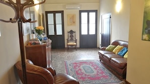 Un salon chaleureux avec un canapé en cuir marron, une table basse en bois et un tapis à motifs. On y trouve une chaise en bois et un bureau agrémenté d’un vase de fleurs. La pièce présente un style traditionnel, avec un plafond en bois et une grande fenêtre.