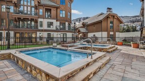 A pool area with two hot tubs, stone-tiled flooring, and a wooden building with balconies in the background. - Slopeside Townhome in Beaver Creek | Chalet Montsor (Beaver Creek)