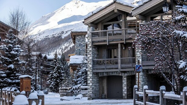 A snow-covered building with a stone facade and wooden balconies, set against a backdrop of snow-capped mountains. - Joli Chalet, Vieux Village (Val-d'Isère)