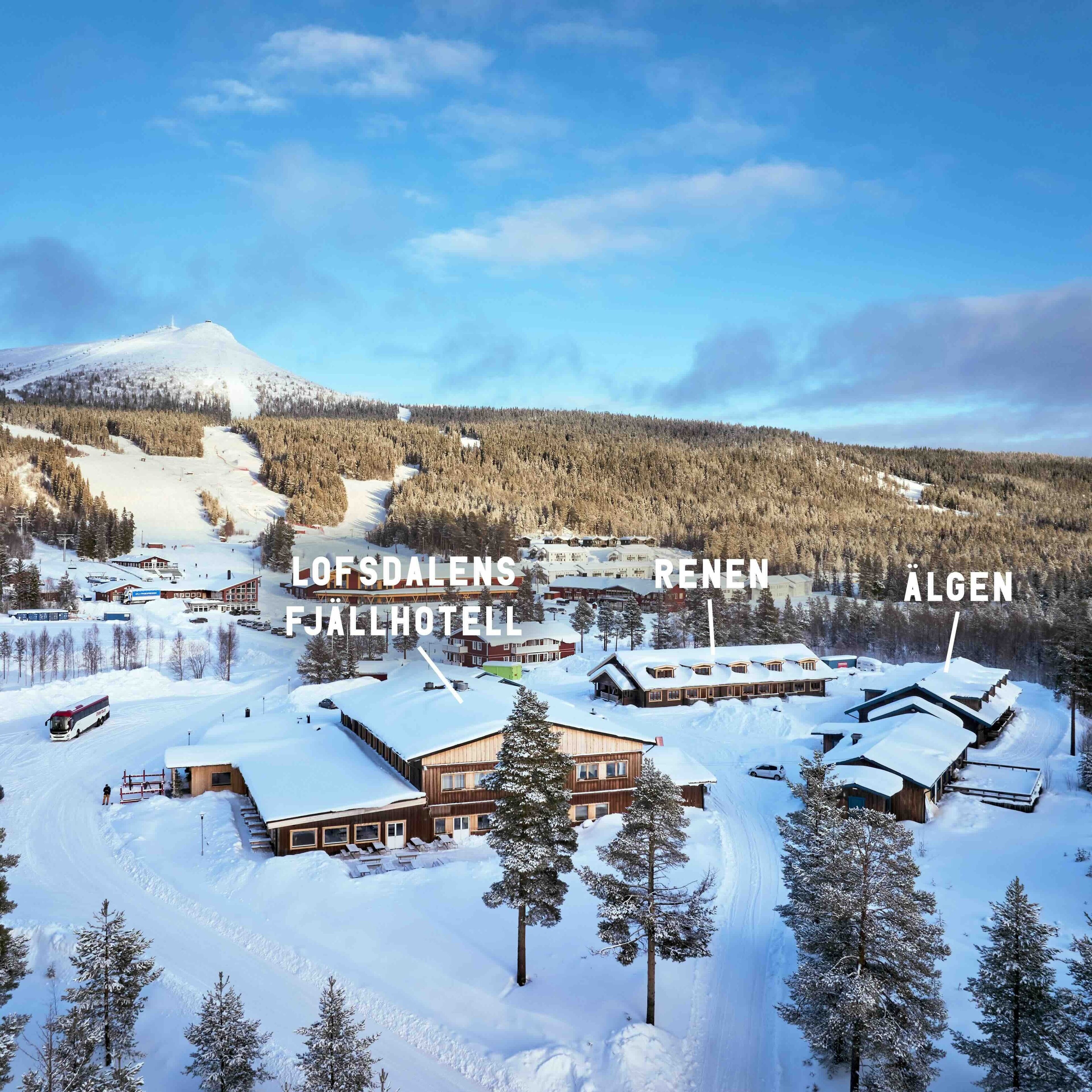 A snow-covered mountain resort with multiple buildings, including a hotel, cabins, and a ski lift.