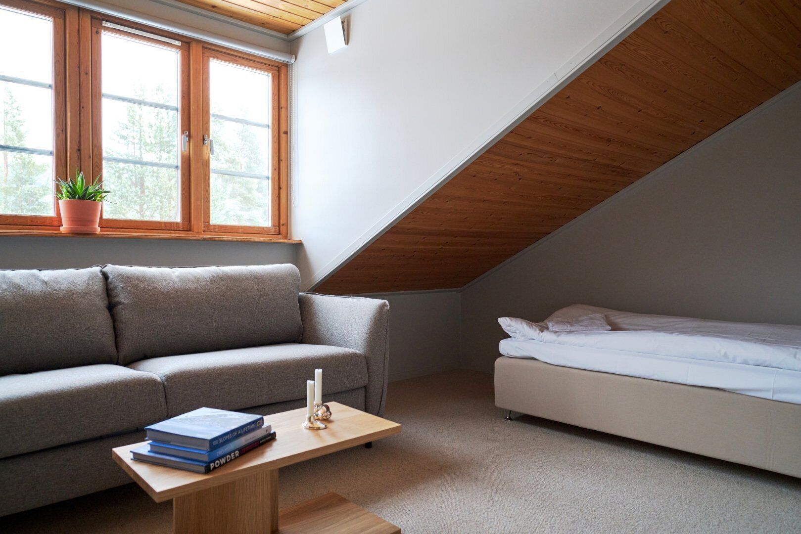 A small, modern room with a sloped ceiling, a grey sofa, a white bed, and a small table with books and a candle.
