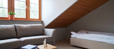 A small, modern room with a sloped ceiling, a grey sofa, a white bed, and a small table with books and a candle.