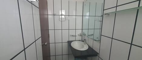 Casa de banho com paredes em azulejos brancos, bancada preta, lavatório branco, cabine de duche em vidro e espelho.