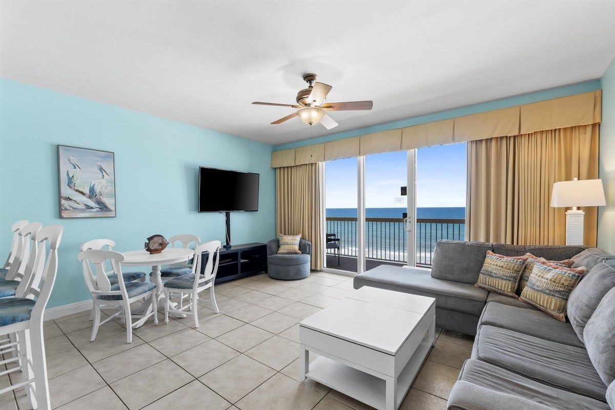 A living room with a balcony, a flat-screen TV, a ceiling fan, and a view of the ocean.