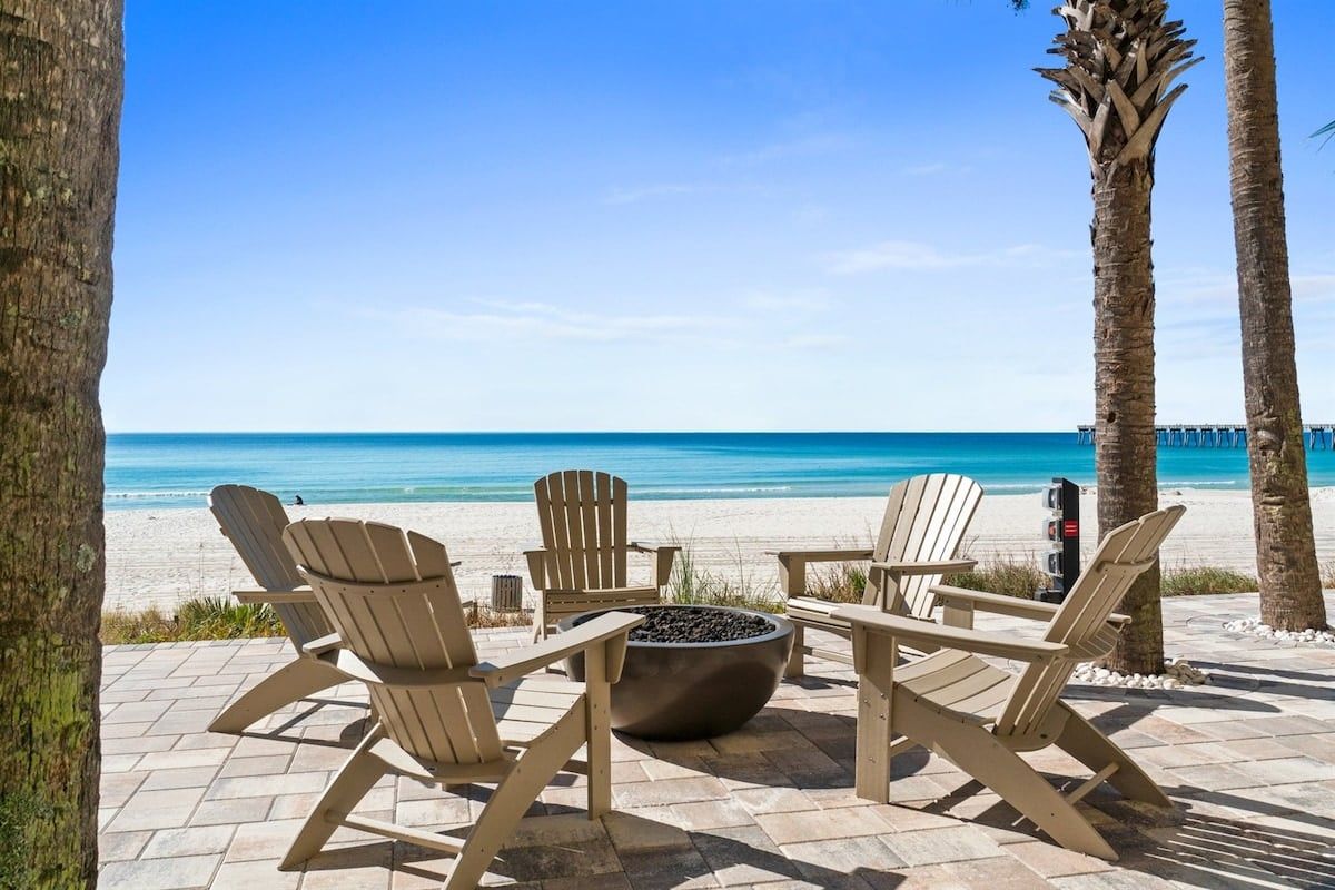 A beachside seating area with Adirondack chairs and a fire pit, overlooking a clear blue ocean.