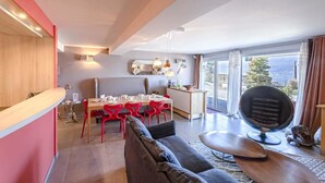 A modern living room with a sofa, a dining area with red chairs, a kitchen with a mirrored backsplash, and a large window offering a view. - Le Clos du Lac: Villa with lake view (Veyrier-du-Lac)