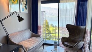 A modern living room with a sofa, armchair, coffee table, and desk. There are paintings on the wall, a window with curtains, and an air conditioning unit. - Le Clos du Lac: Villa with lake view (Veyrier-du-Lac)