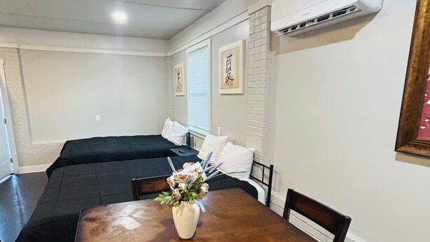 A hotel room with a wooden dining table, a single chair, a small vase with flowers, a bed with a black mattress, and a wall-mounted air conditioner. - Madison gate studio (Goldsboro)