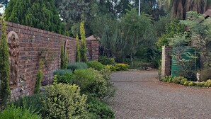 A gravel pathway leading through a garden with a brick wall and statues, surrounded by various plants and trees. - Flower Lane Guesthouse  (Kathu)