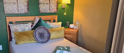Una cama bien tendida con una colcha verde y blanca, un cabecero de madera y almohadones decorativos. Hay una lámpara de pared y una obra de arte enmarcada sobre la cama.