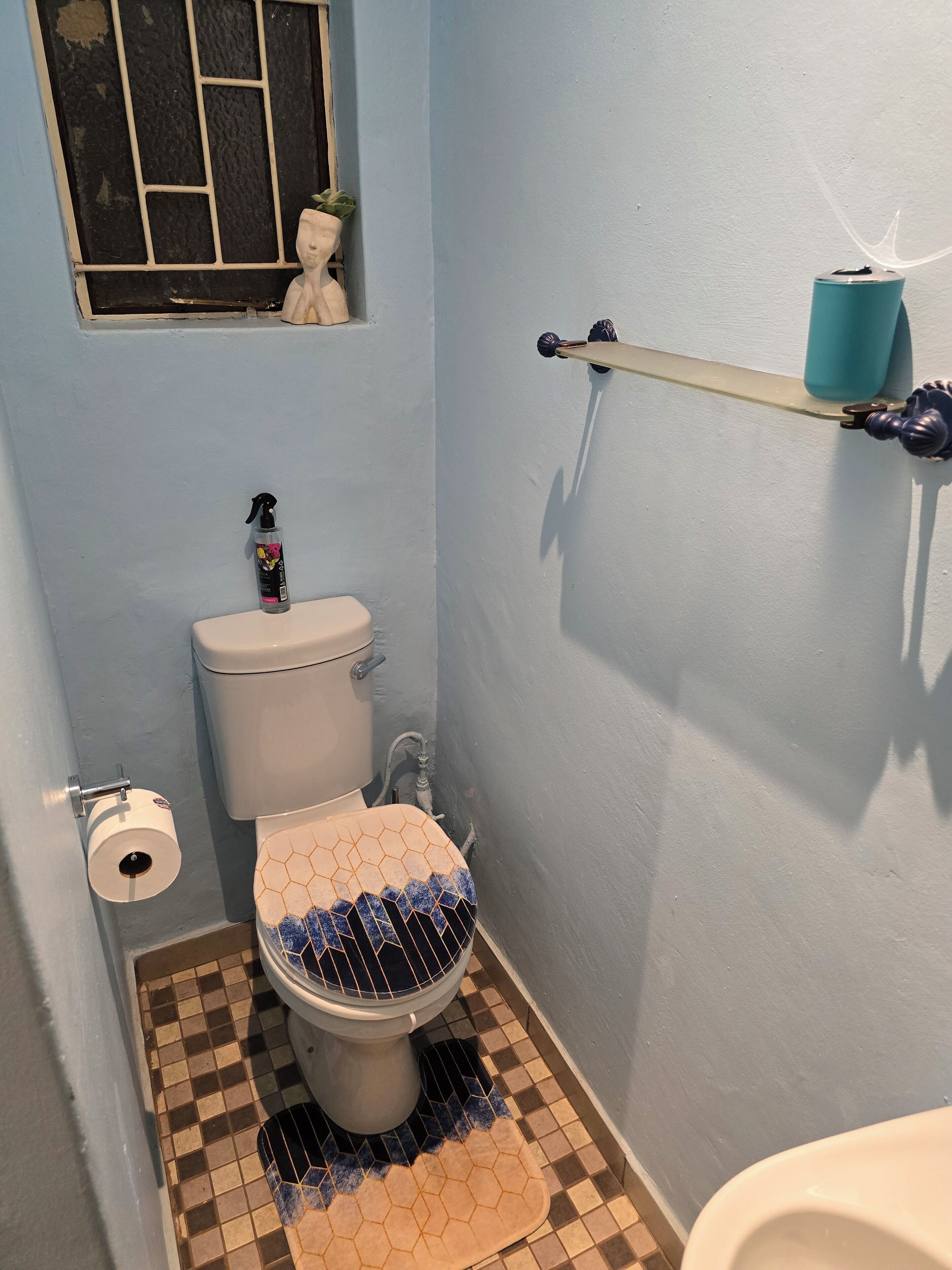 A small bathroom with a toilet, a window with bars, a shelf with a blue container, and a tiled floor.