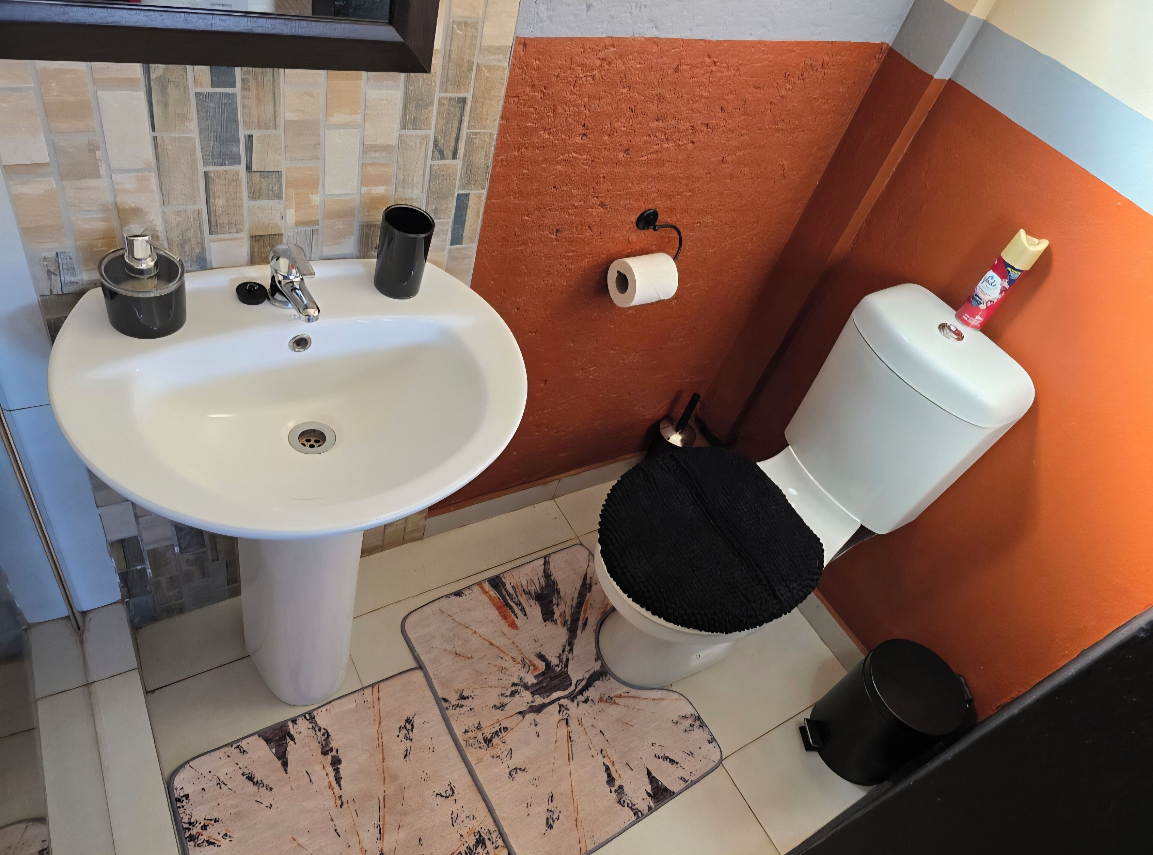 A bathroom with a white sink, a toilet with a black seat cover, a roll of toilet paper, and a trash bin.
