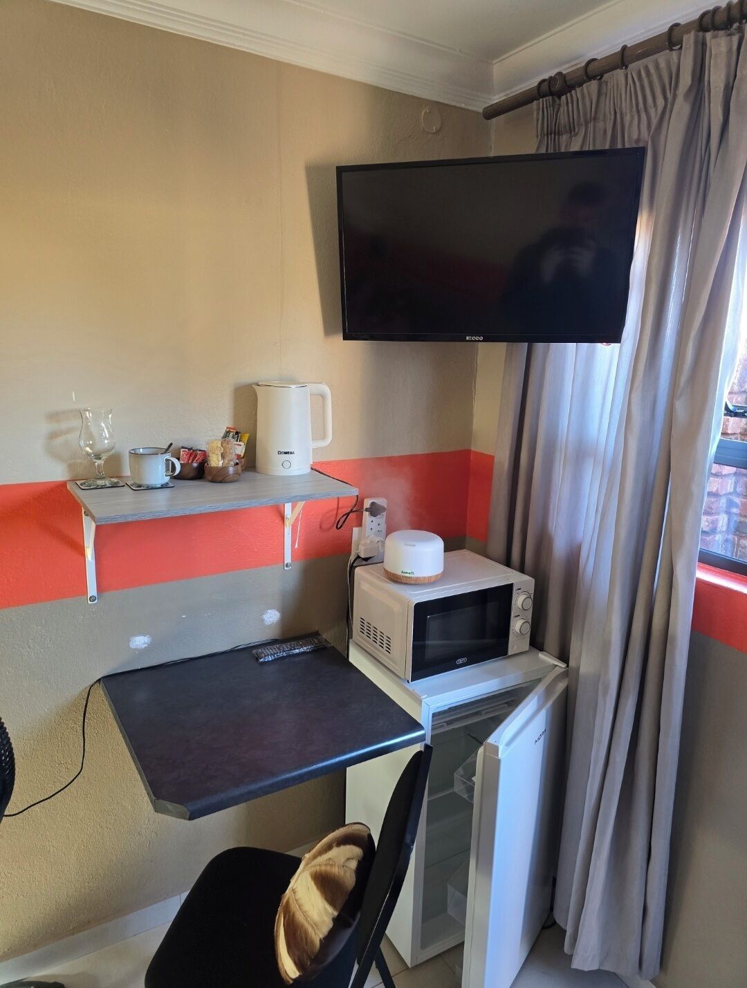 Ett kompakt hotellrum med ett litet pentry, en platt-TV som är monterad på väggen och ett fönster med gardiner.