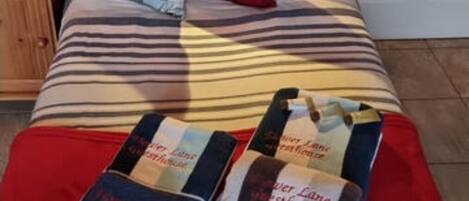 Een opgemaakt bed met een gestreept dekbed, een rood kussen en meerdere kussens met een blauw-wit patroon. Er staat een nachtkastje met boeken en een lamp.