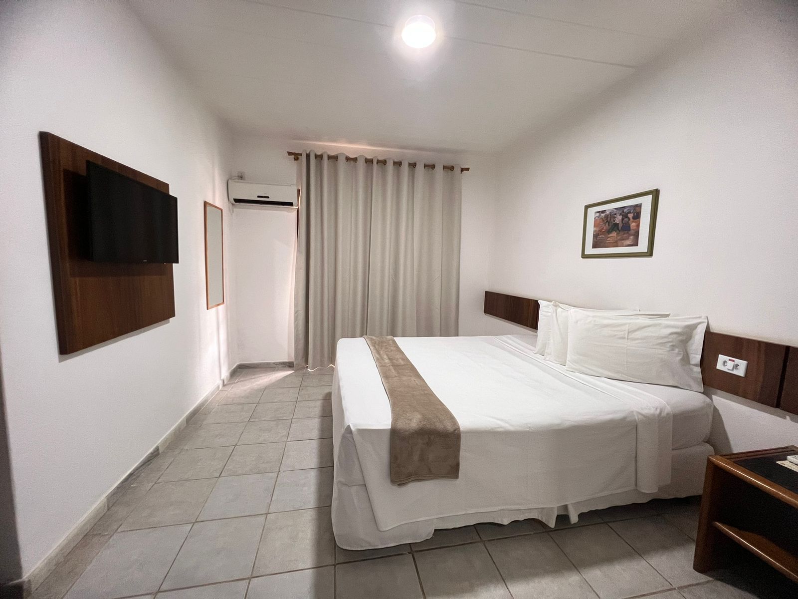 Quarto de hotel com cama, televisão fixada na parede, quadro emoldurado e janela com cortinas.