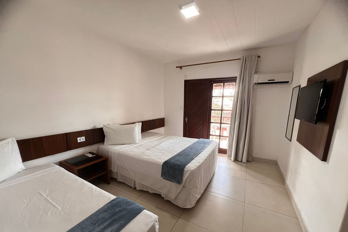 Une chambre d’hôtel avec deux lits, une tête de lit en bois, une table de chevet, un téléviseur fixé au mur et une fenêtre avec des rideaux.