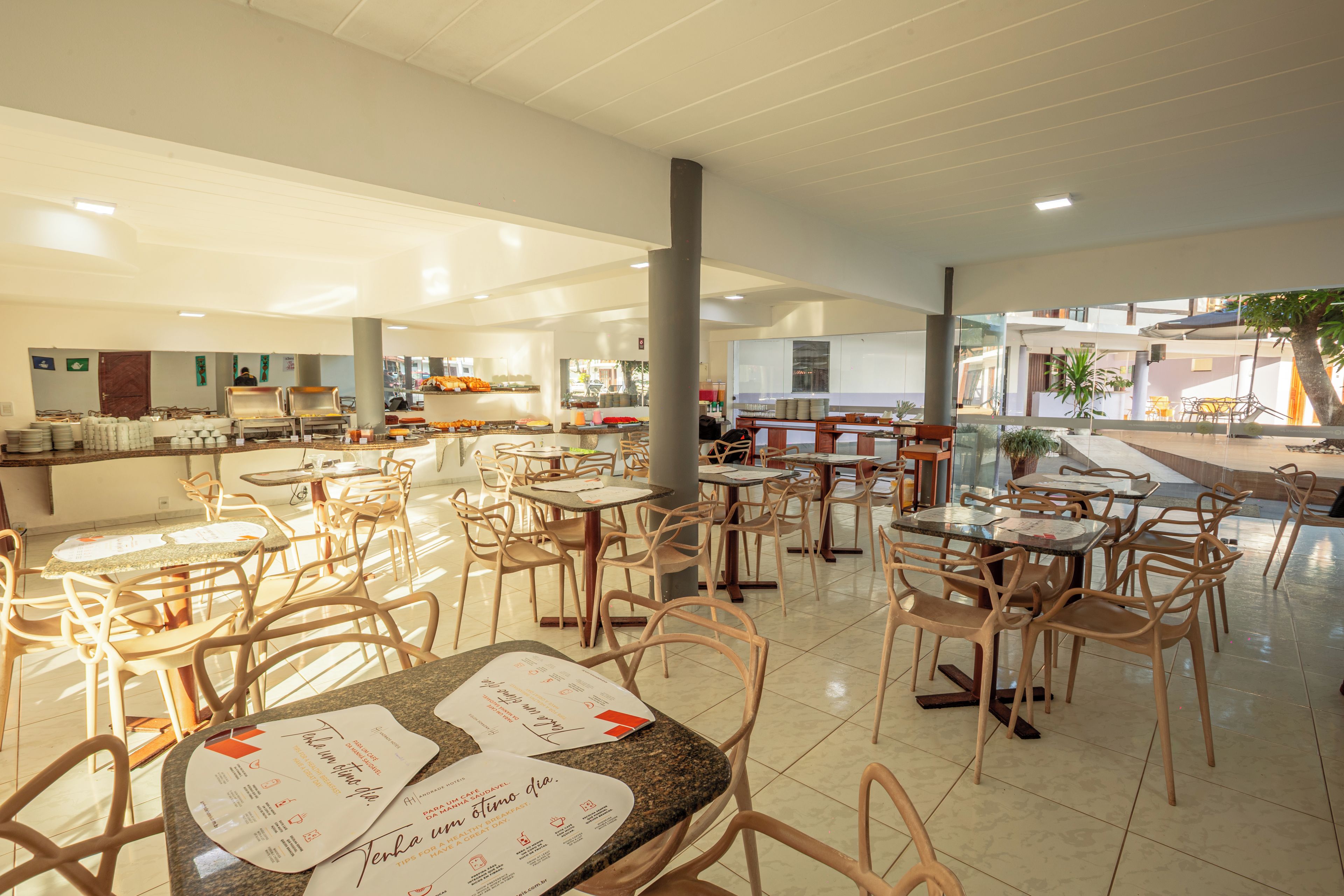 Uma ampla área de refeições interna com várias mesas e cadeiras, uma cozinha ao fundo e vista para o exterior através de grandes janelas.