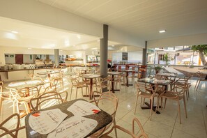 Un vaste espace de restauration intérieur, doté de nombreuses tables et chaises, avec un coin cuisine à l’arrière-plan et une vue sur l’extérieur grâce à de grandes fenêtres.