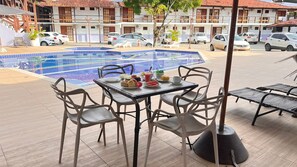 Uma área de refeições à beira da piscina, com uma mesa posta para quatro pessoas, rodeada de cadeiras, e uma piscina ao fundo.