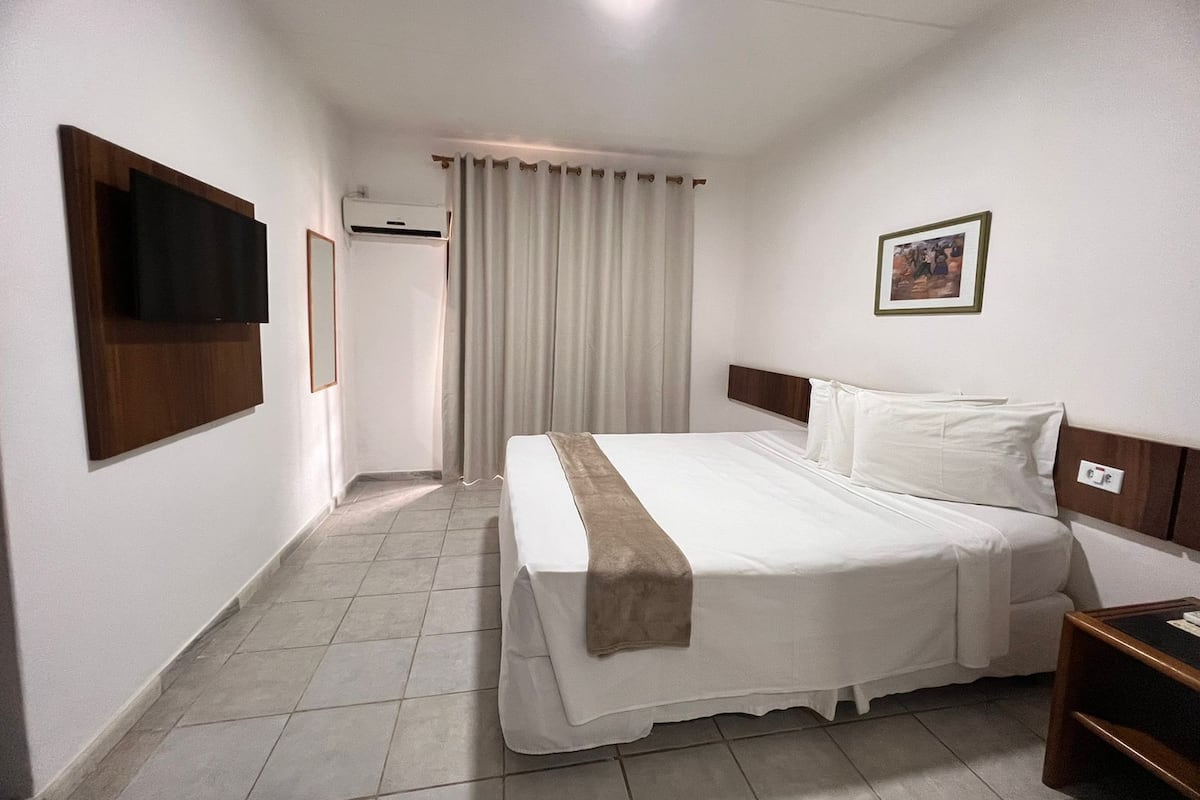 A hotel room with a bed, a TV mounted on the wall, a framed picture, and a window with curtains.