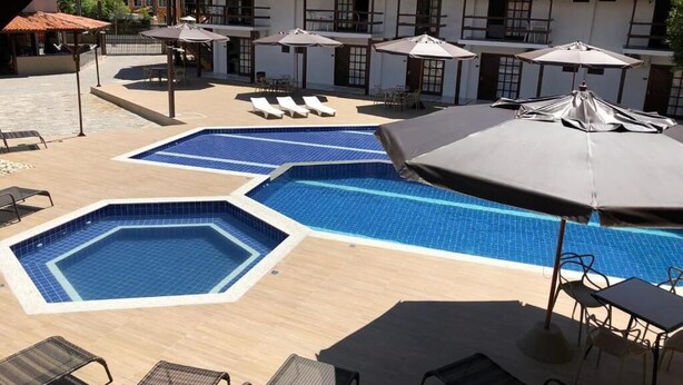 A pool area with multiple pools, lounge chairs, umbrellas, and a building with a tiled roof in the background. - Rede Andrade Estrela Dalva Praia (Porto Seguro)