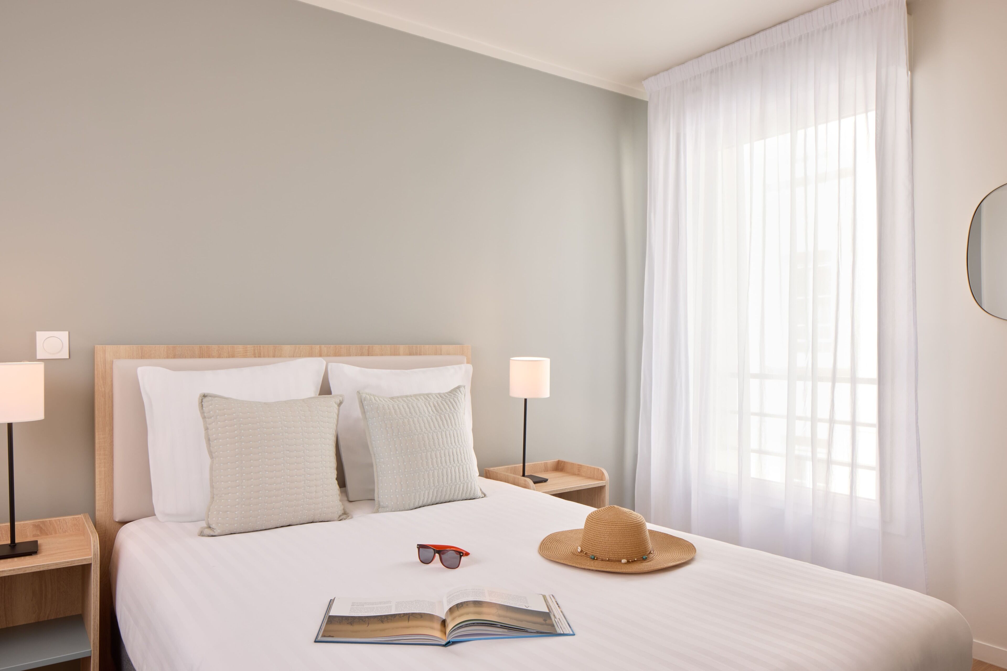 Una cama bien hecha con almohadas, un sombrero, gafas de sol y un libro abierto en una habitación limpia y de tonos claros, con cabecera de madera y lámparas de noche.