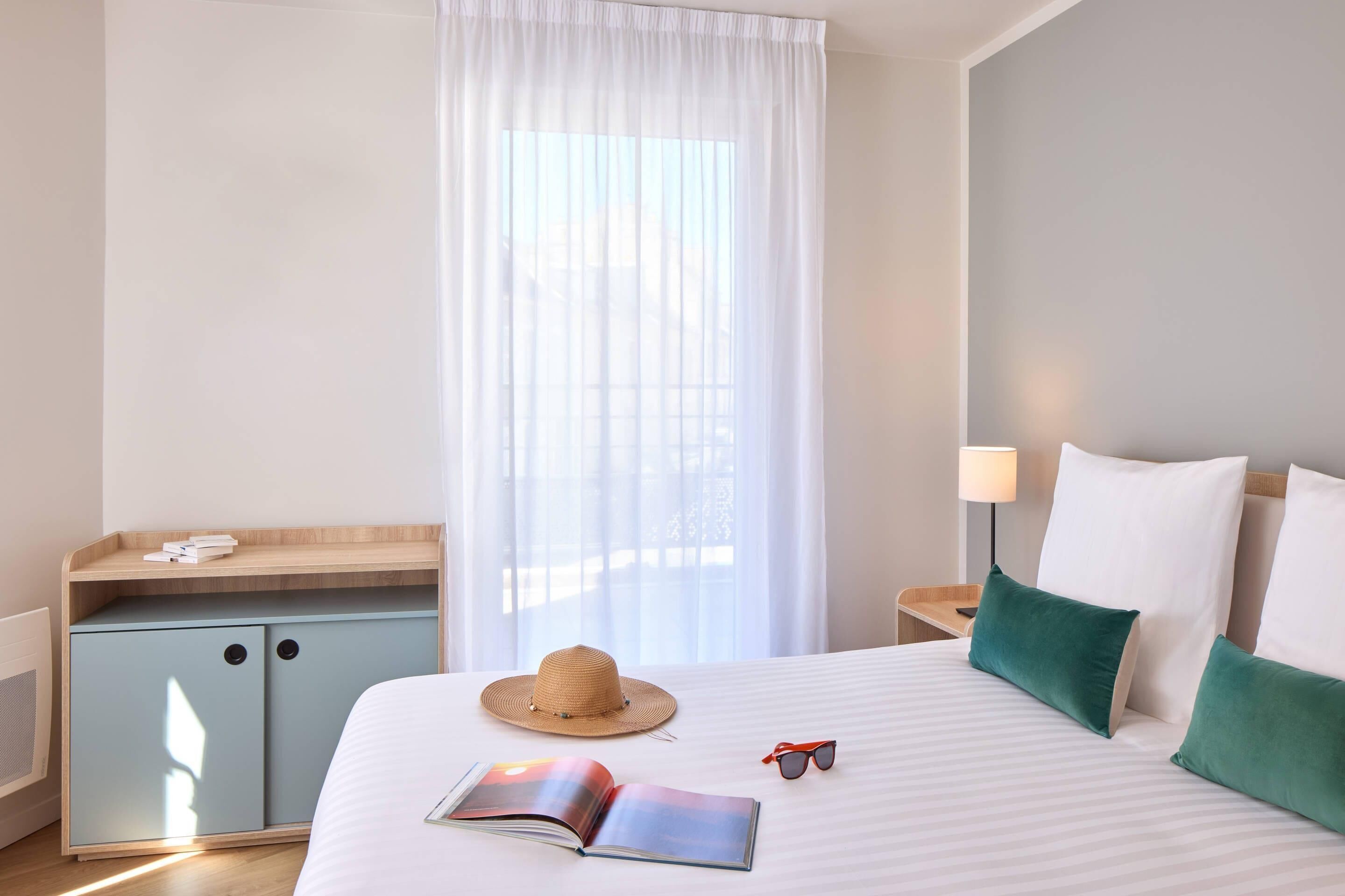 Een hotelkamer met een bed, nachtkastje, bureau en een raam met dunne gordijnen.