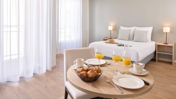 Una habitación de hotel con una cama bien hecha, una mesa de desayuno con croissants, zumo de naranja y una taza de café.