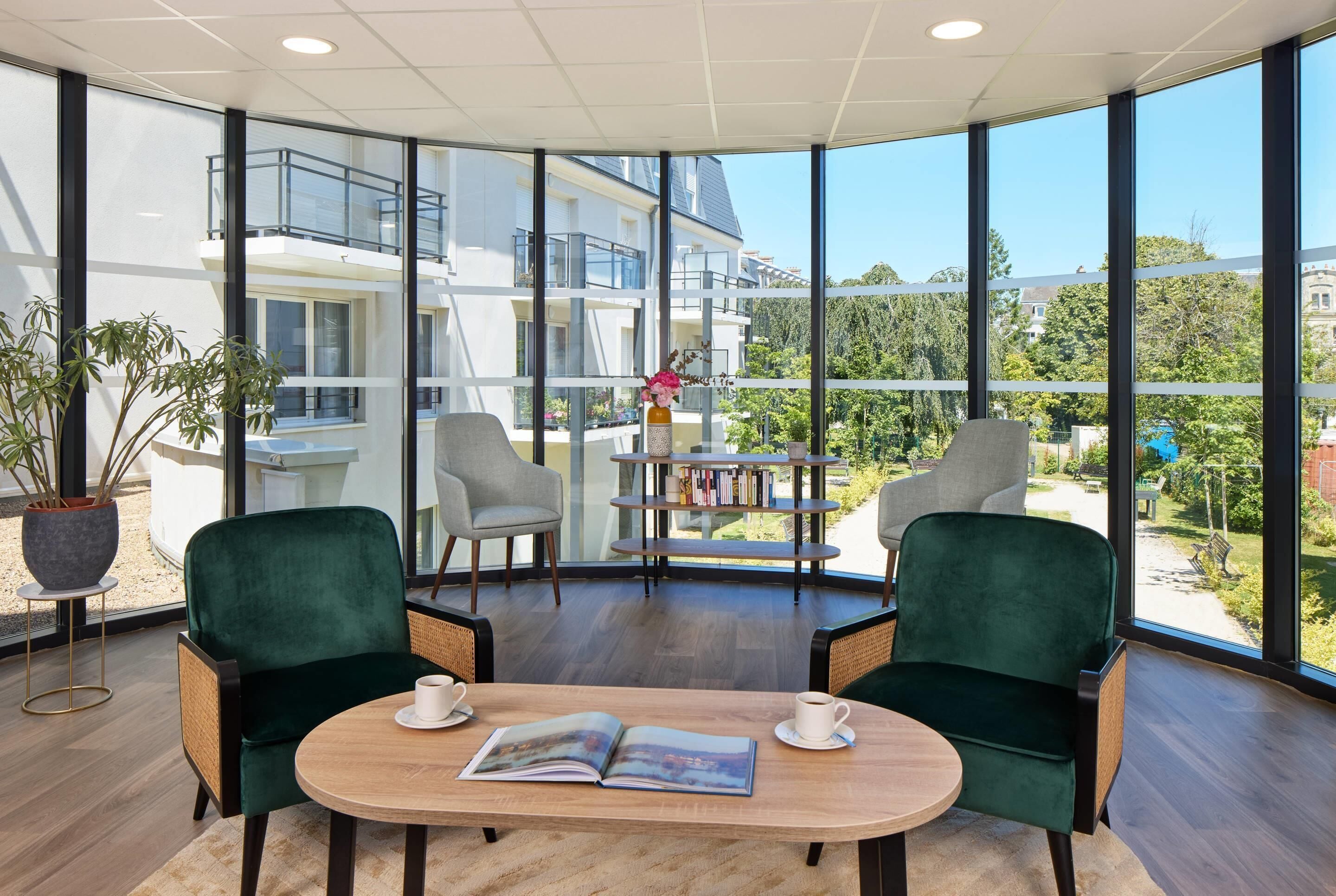 A modern room with large windows, two green chairs, a wooden table, and a bookshelf.