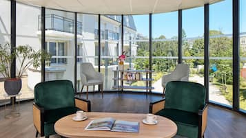 A modern room with large windows, two green chairs, a wooden table, and a bookshelf.
