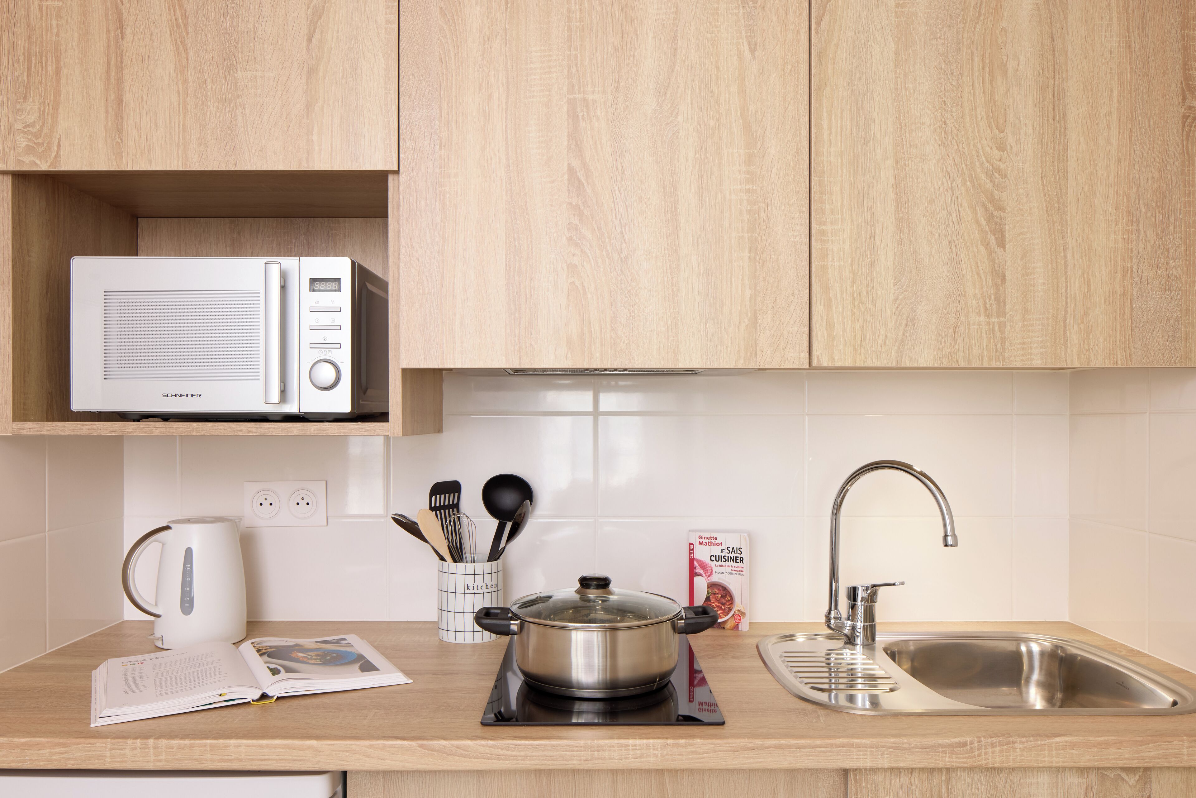 A kitchen with wooden cabinets, a microwave, a kettle, a pot on an induction cooktop, a cookbook, and a stainless steel sink.