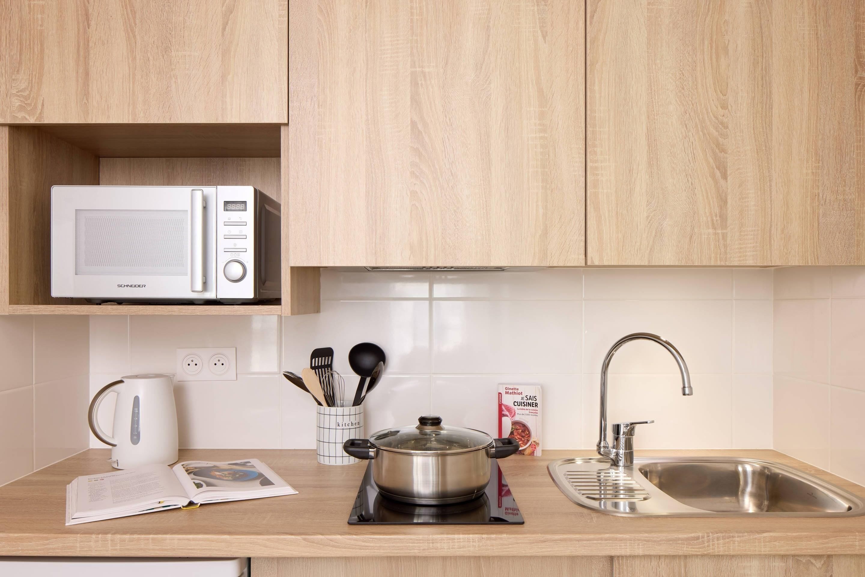 A kitchen with a microwave, a kettle, a pot on a portable stove, a sink, and a magazine on the counter.