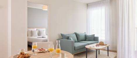 A modern hotel room with a round dining table set for breakfast, a sofa, and a small side table with a hat.