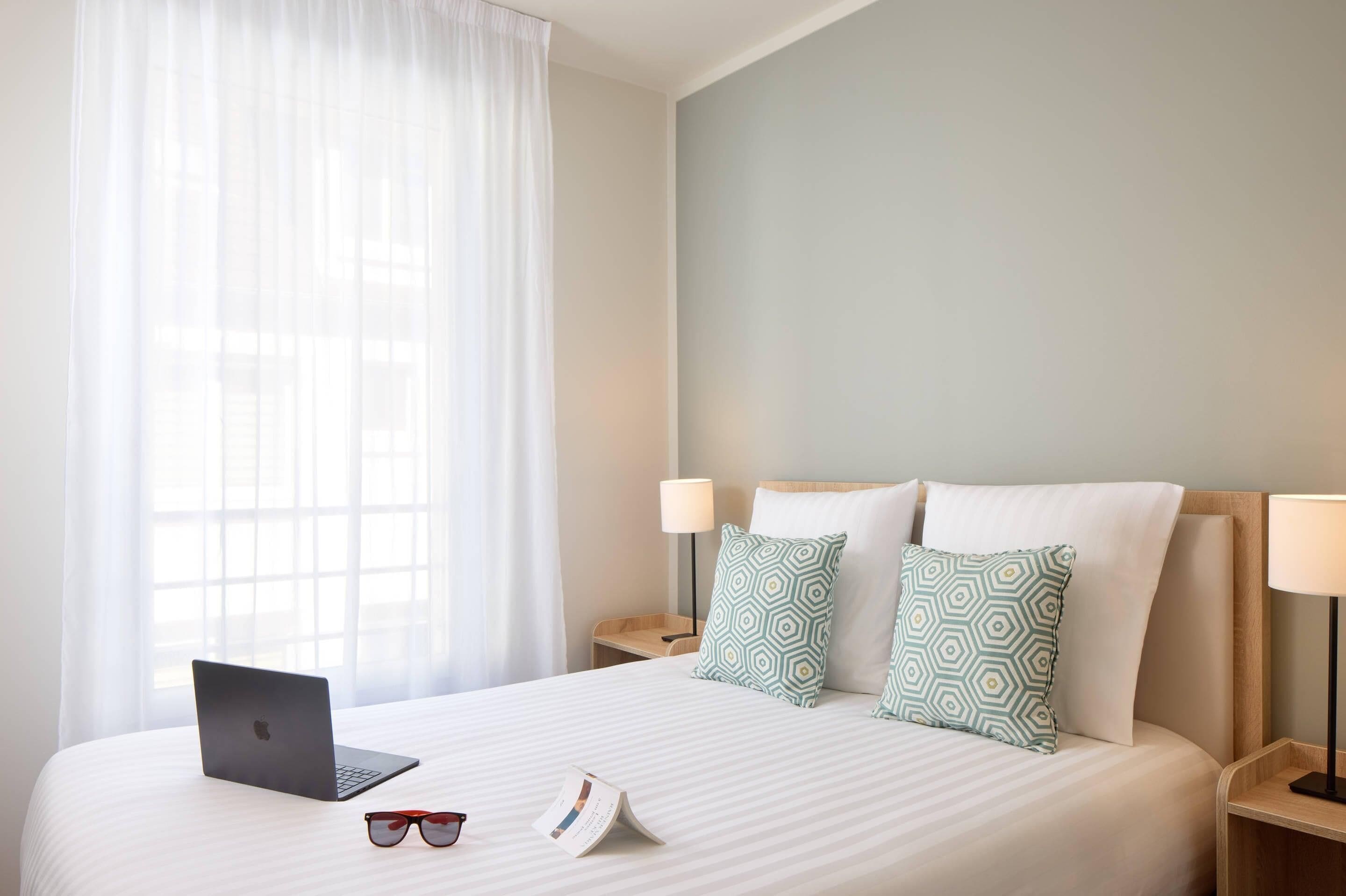 A neatly made bed with a laptop, sunglasses, and a brochure on it. There are two bedside lamps and patterned pillows.