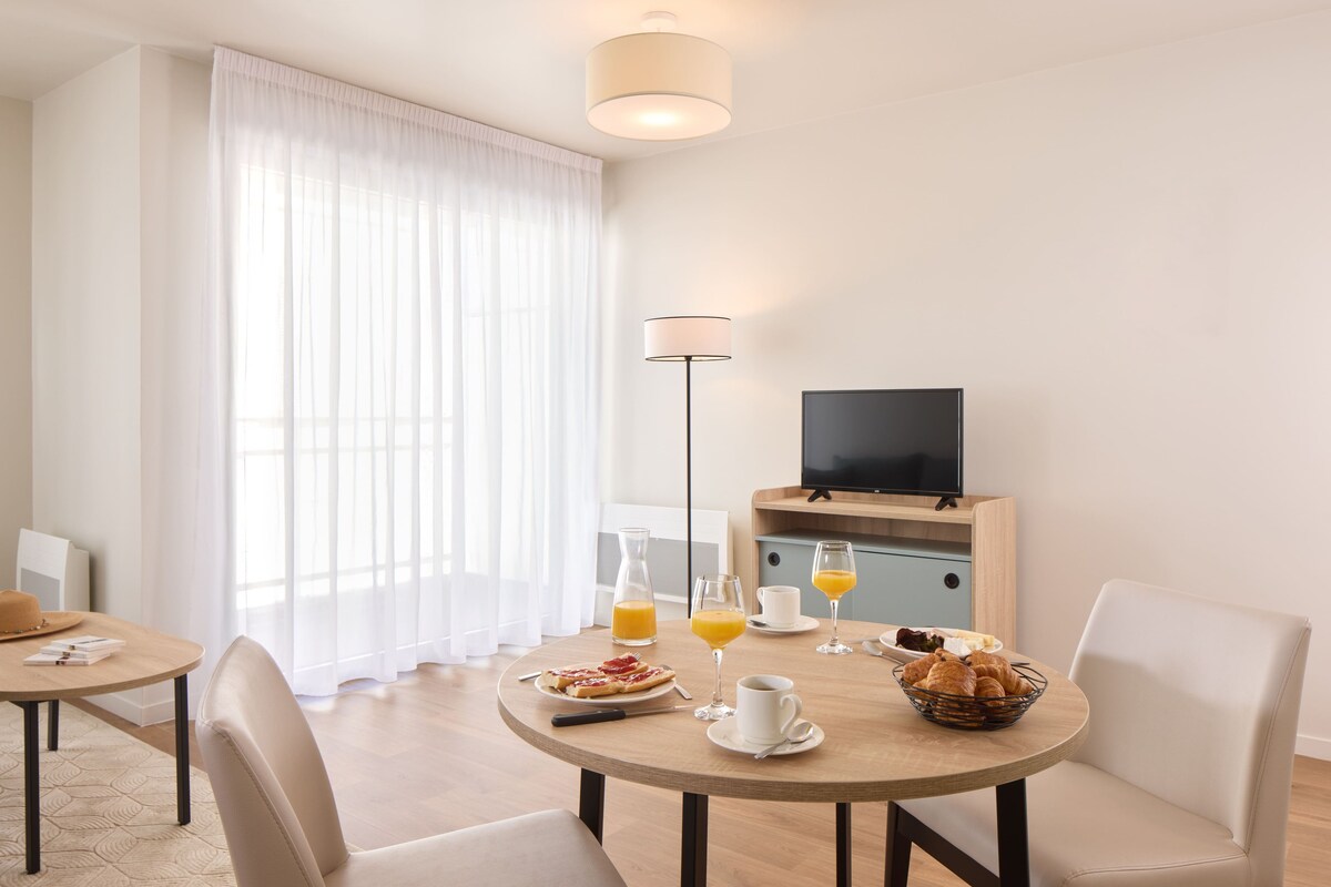 Un área de comedor con una mesa dispuesta para el desayuno, un televisor y un amplio ventanal con cortinas traslúcidas.