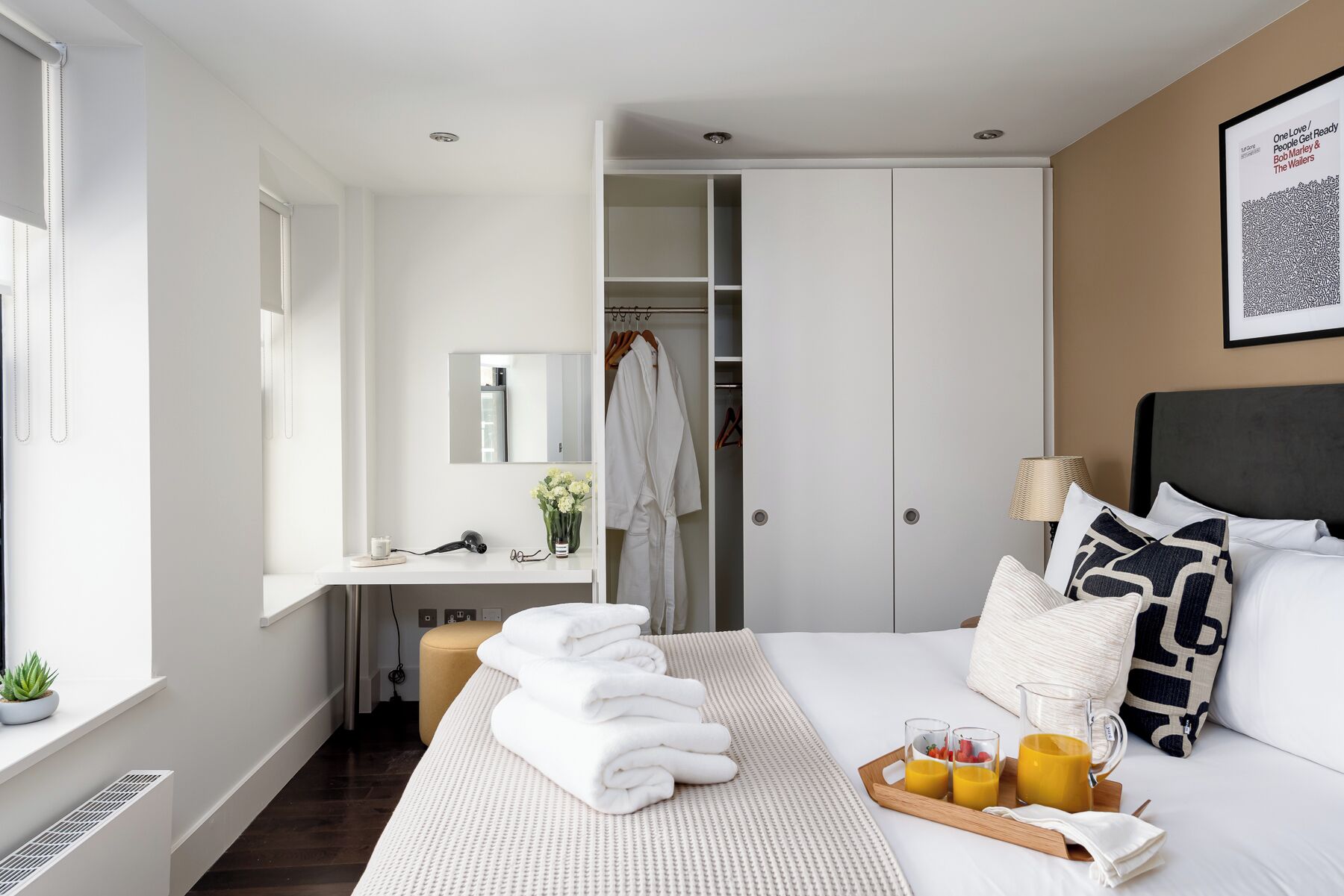 A modern hotel room with a neatly made bed, a bedside table with a lamp, a tray with breakfast, and a closet with hanging clothes.