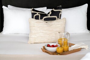 Un letto rifatto con lenzuola bianche, un cuscino decorativo in tessuto intrecciato, una ciotola di fragole e un vassoio con bicchieri di succo d'arancia.