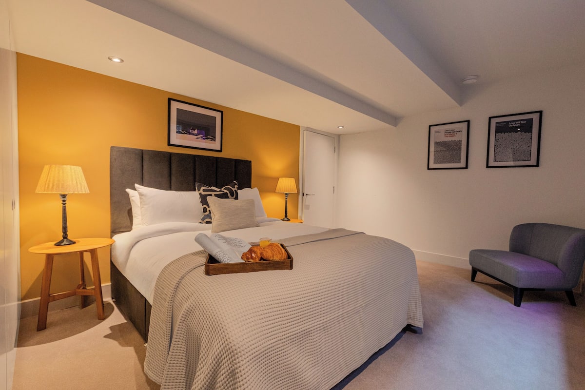 A hotel room with a large bed, two bedside lamps, a chair, and a tray with croissants.