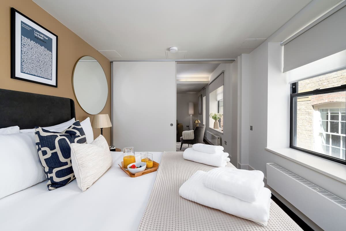 A modern hotel room with a large bed, a bedside table with a tray of breakfast, a large mirror, and a view of the outside.