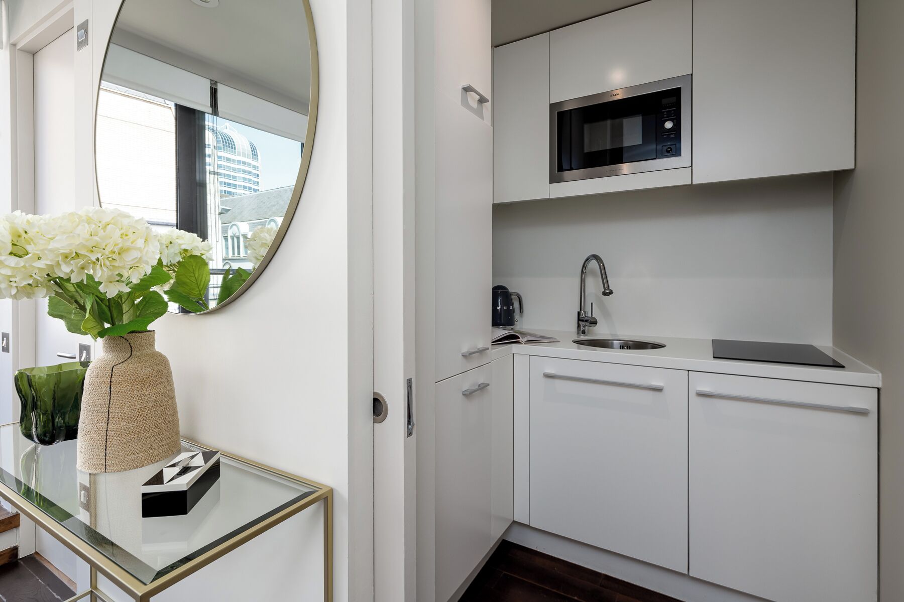 A modern kitchen with white cabinets, a stainless steel sink, a microwave, and a glass coffee table with a vase of flowers.