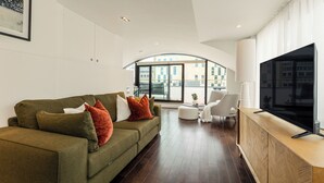 A modern living room with a green sofa, a flat-screen TV, and a view of the city through large windows. - Your Apartment - Tower of London (London)