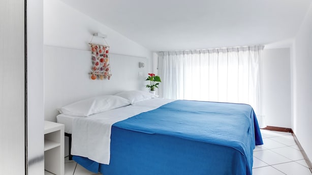 A bedroom with a bed, white walls, a window with curtains, a bedside table, and a potted plant. - Three-room flat Apartment Scola 21 (Gabicce Mare)
