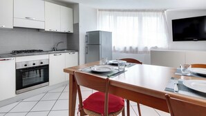 A modern kitchen with white cabinets, a built-in oven, a gas stove, and a refrigerator. There is a dining table with chairs and a TV mounted on the wall. - Three-room flat Apartment Scola 21 (Gabicce Mare)