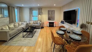 A modern living room with a sofa, coffee table, dining table set for four, and a flat-screen TV mounted on the wall.