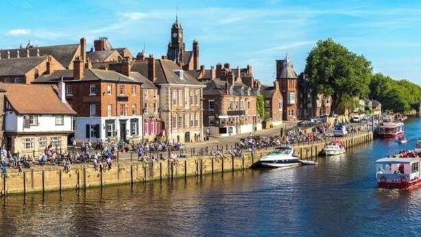 A riverside town with historic buildings, a prominent clock tower, and a promenade with people and boats.