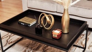 A black coffee table with a Chanel perfume, a black box, a gold ring, a gold vase, and a lit candle.