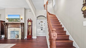 A grand staircase with wooden railings leading to a landing, a fireplace with a mantel, and a large framed picture on the wall.