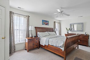 A bedroom with a wooden bed, bedside tables, a ceiling fan, a window with curtains, a painting, and a mirror.