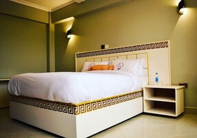 Una cama con cabecera blanca y un diseño decorativo, una mesita de noche con una botella, y una lámpara fijada a la pared.
